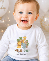 A smiling baby boy wearing a white long-sleeved shirt with a jungle-themed graphic and the text "WILD ONE Oliver" printed on it.
