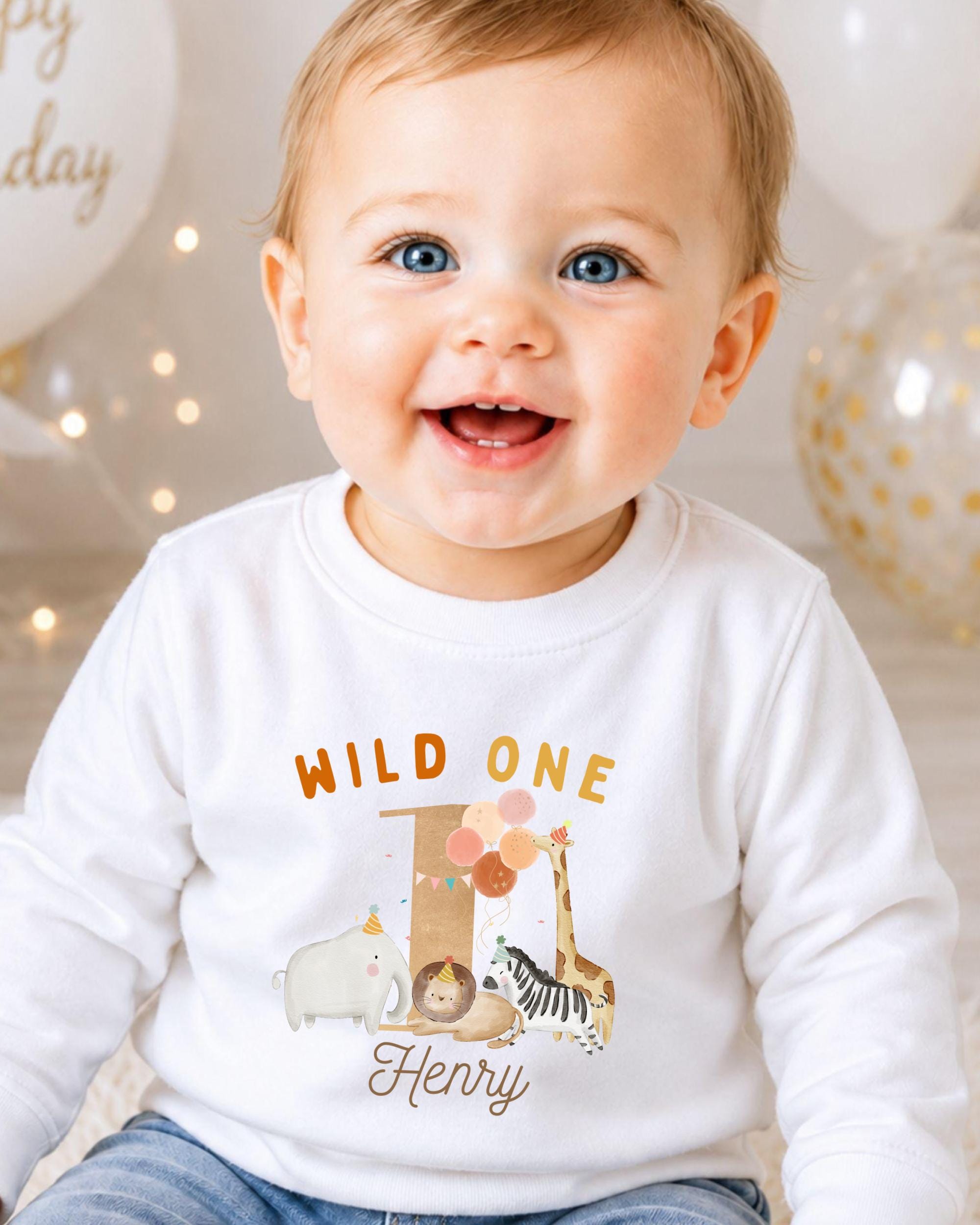 A smiling baby boy wearing a white shirt with a cartoon animal and the text "Wild One Henry" is the focus of the image.