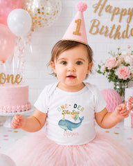 A young girl wearing a birthday hat and a white t-shirt with a cartoon whale is posing in front of a birthday cake and balloons.