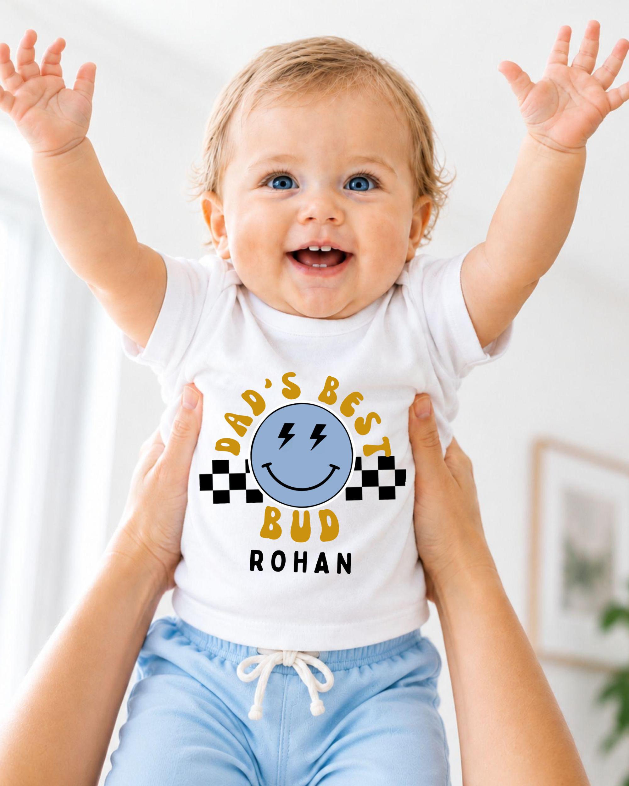 A smiling baby wearing a white t-shirt with a graphic design and text, being held up by someone's arms.