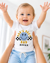 A smiling baby wearing a white t-shirt with a graphic design and text, being held up by someone's arms.