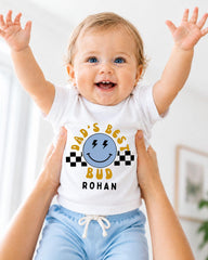 A smiling baby wearing a white t-shirt with a graphic design and text, being held up by someone's arms.