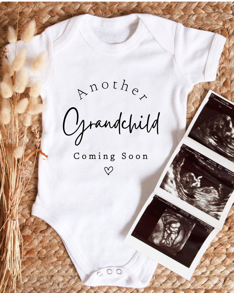 A white baby bodysuit with the text "Another Grandchild Coming Soon" printed on it, along with a heart symbol. Next to the bodysuit is a set of three black and white baby ultrasound images.