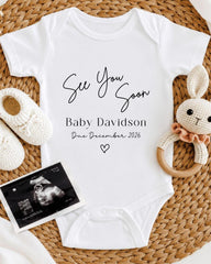 a white baby onesie with the text "See You Soon Baby Davidson Due December 2026" printed on it, along with a crocheted bunny toy and a black and white photograph of an infant's head.