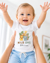 A smiling baby wearing a white t-shirt with a jungle-themed design and the text "WILD ONE Oliver" is being held up by someone's arms.