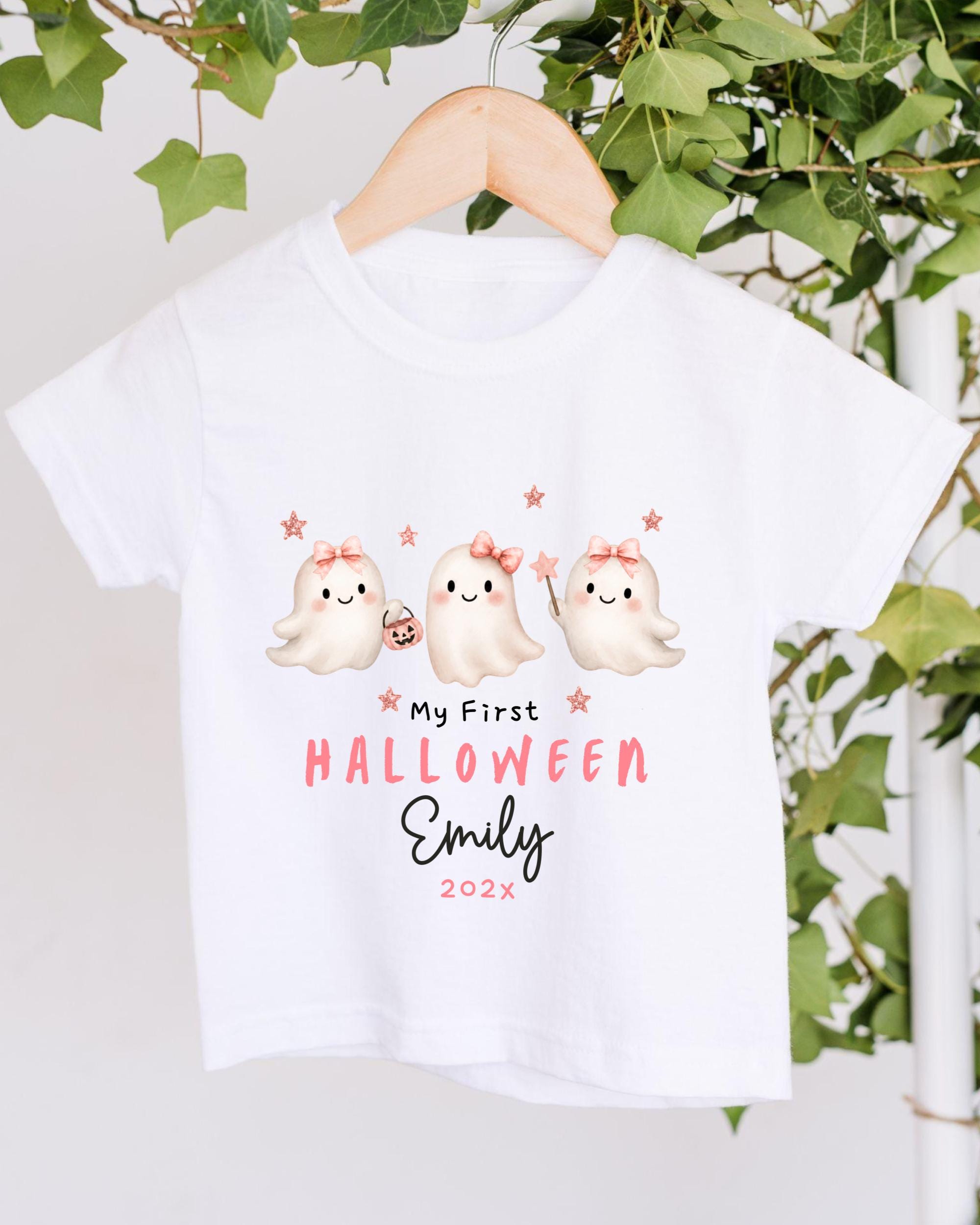 A white t-shirt with a Halloween-themed design featuring three cute ghost characters with bows and stars. The text on the t-shirt reads "My First Halloween Emily 2023".