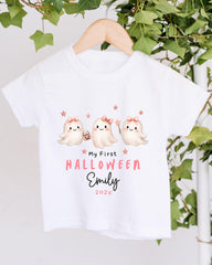 A white t-shirt with a Halloween-themed design featuring three cute ghost characters with bows and stars. The text on the t-shirt reads "My First Halloween Emily 2023".
