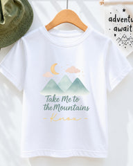 A white t-shirt with a graphic design of mountains and a crescent moon, along with the text "Take Me to the Mountains" and "Knox" printed on it.