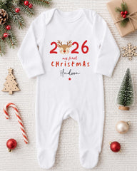 A white onesie with the text "26 my first christmas Hudson" printed on it, surrounded by Christmas decorations such as a pine tree, ornaments, and a candy cane.