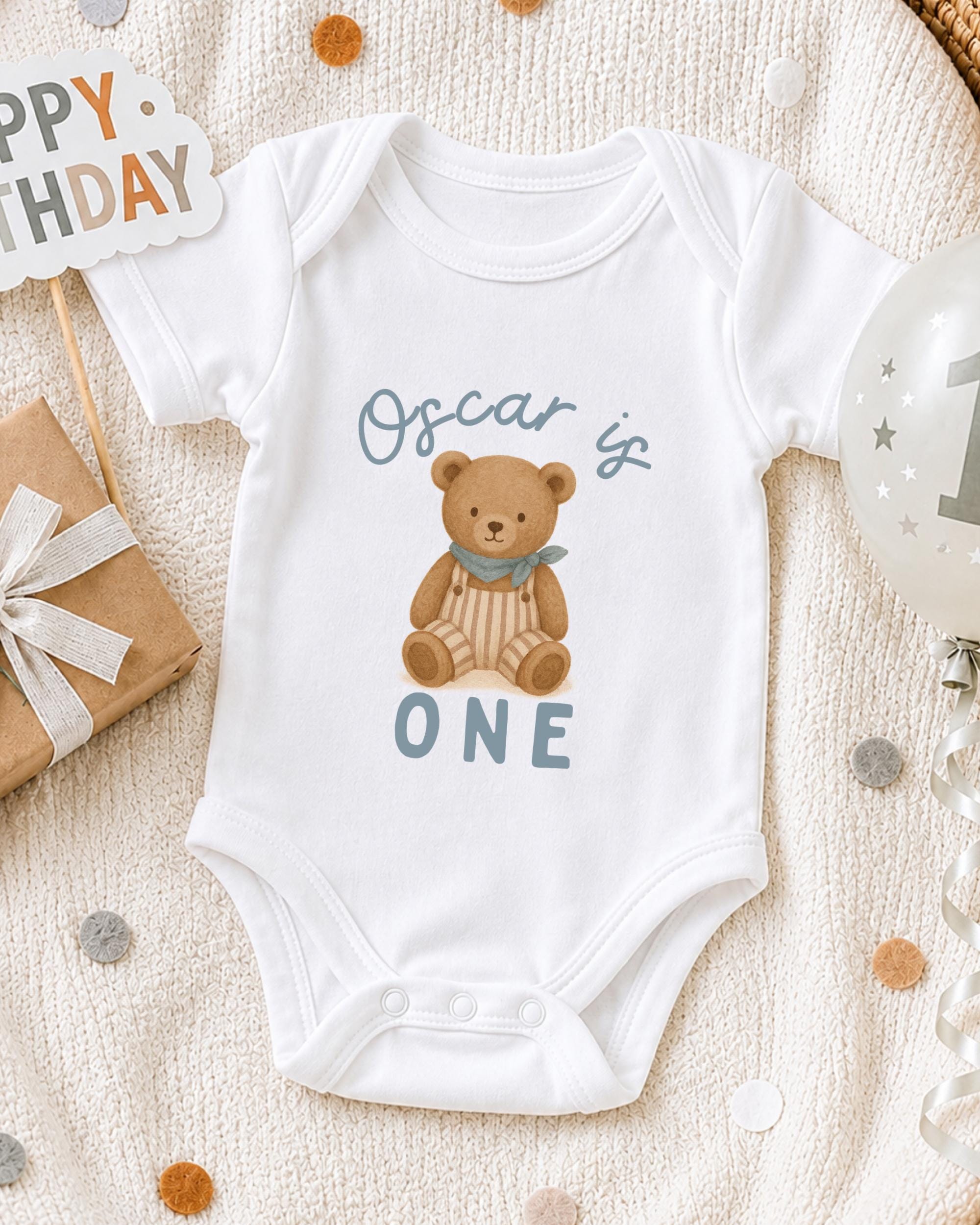 A white onesie with a teddy bear design and the text "Oscar is ONE" printed on it, along with a "Happy Birthday" sign and a birthday balloon.