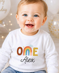 A smiling baby boy wearing a white shirt with a rainbow graphic and the name "Olive" printed on it.