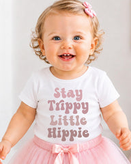 A smiling baby girl wearing a white t-shirt with the text "Stay Trippy Little Hippie" and a pink tutu skirt.