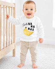 A smiling baby boy wearing a white sweatshirt with a banana graphic and the text "Totally BANANA" is standing next to a wooden crib.