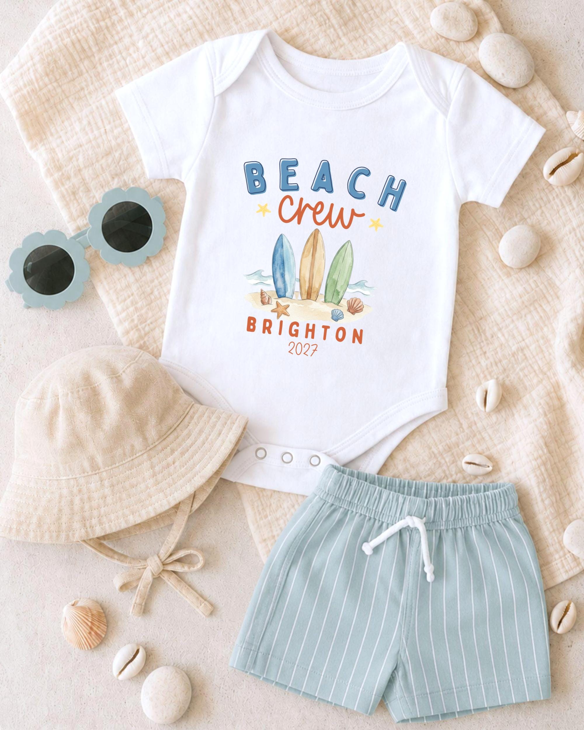 A baby's outfit, including a white onesie with a beach crew design, a beige bucket hat, and striped shorts, along with sunglasses and seashells, is laid out on a beige surface.