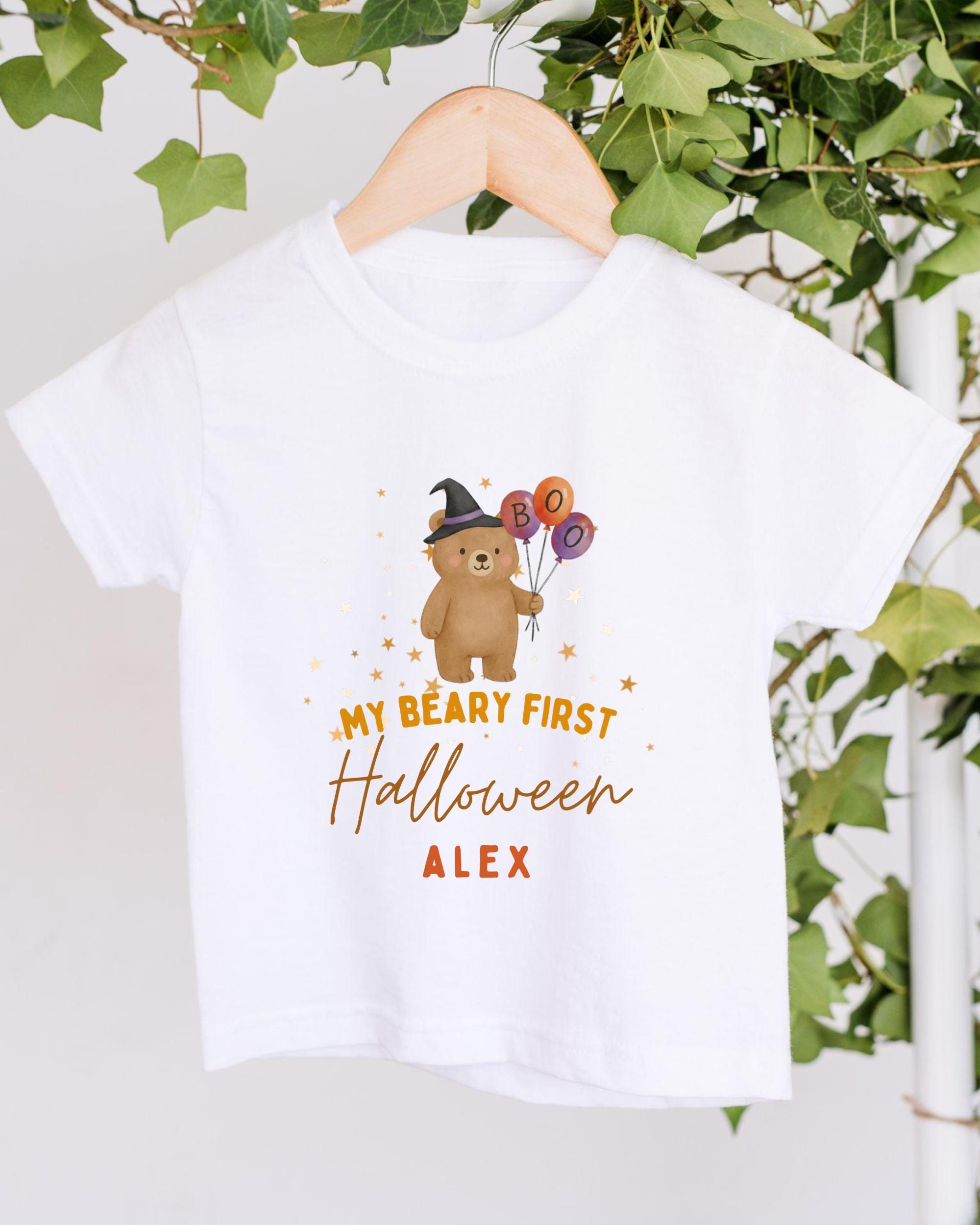 A white t-shirt with a cartoon bear wearing a witch's hat and holding balloons, along with the text "My bear first Halloween Alex" printed on it.