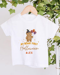 A white t-shirt with a cartoon bear wearing a witch's hat and holding balloons, along with the text "My bear first Halloween Alex" printed on it.