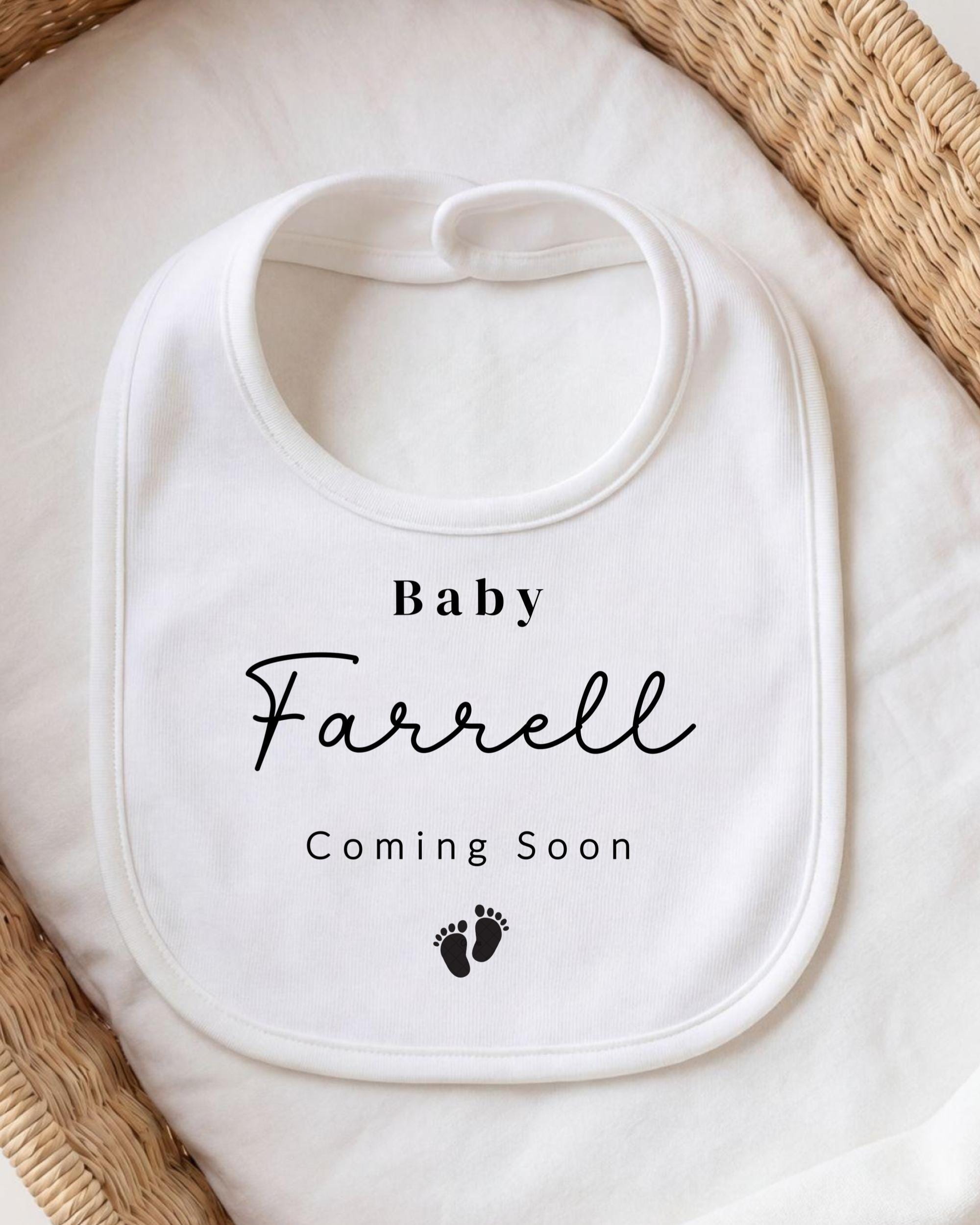 A white bib with the text "Baby Farrell Coming Soon" printed on it, along with a small black footprint icon.
