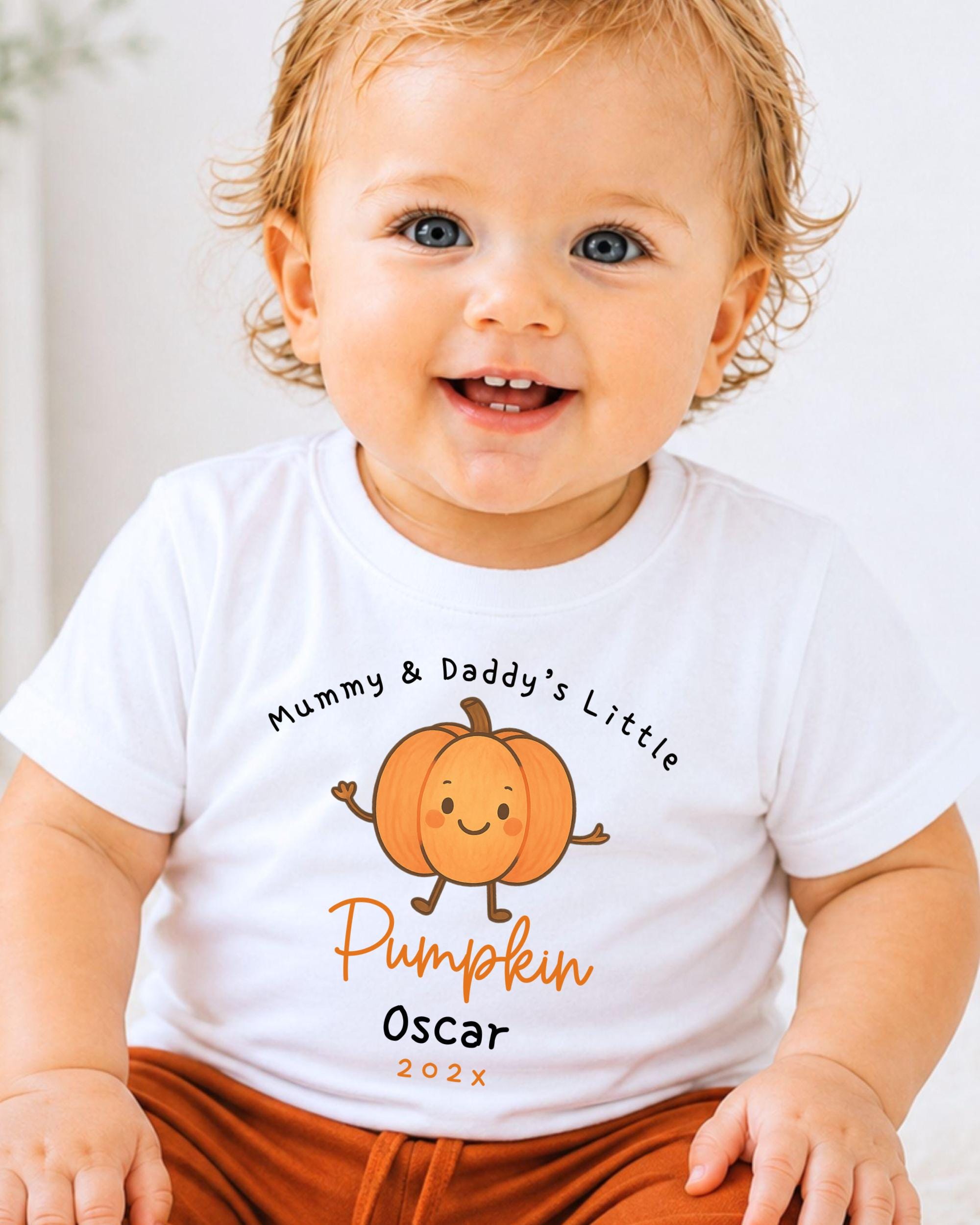 A smiling baby wearing a white t-shirt with a pumpkin graphic and text that says "Mummy & Daddy's Little Pumpkin Oscar 202x".