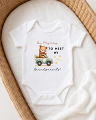 A white onesie with a cartoon bear and the text "On My Way to Meet My Grandparents!" printed on it, placed on a white blanket.