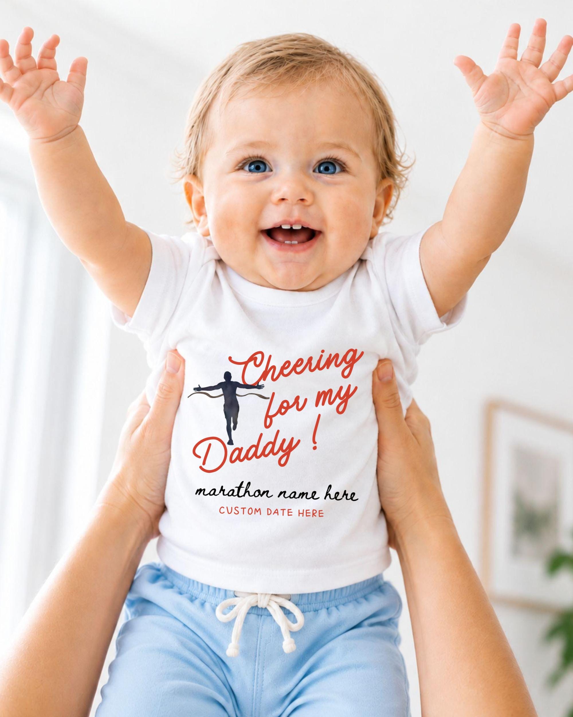 A smiling baby wearing a white t-shirt with a cheerleading graphic and text, being held up by someone's arms.