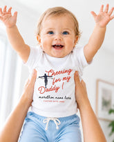A smiling baby wearing a white t-shirt with a cheerleading graphic and text, being held up by someone's arms.