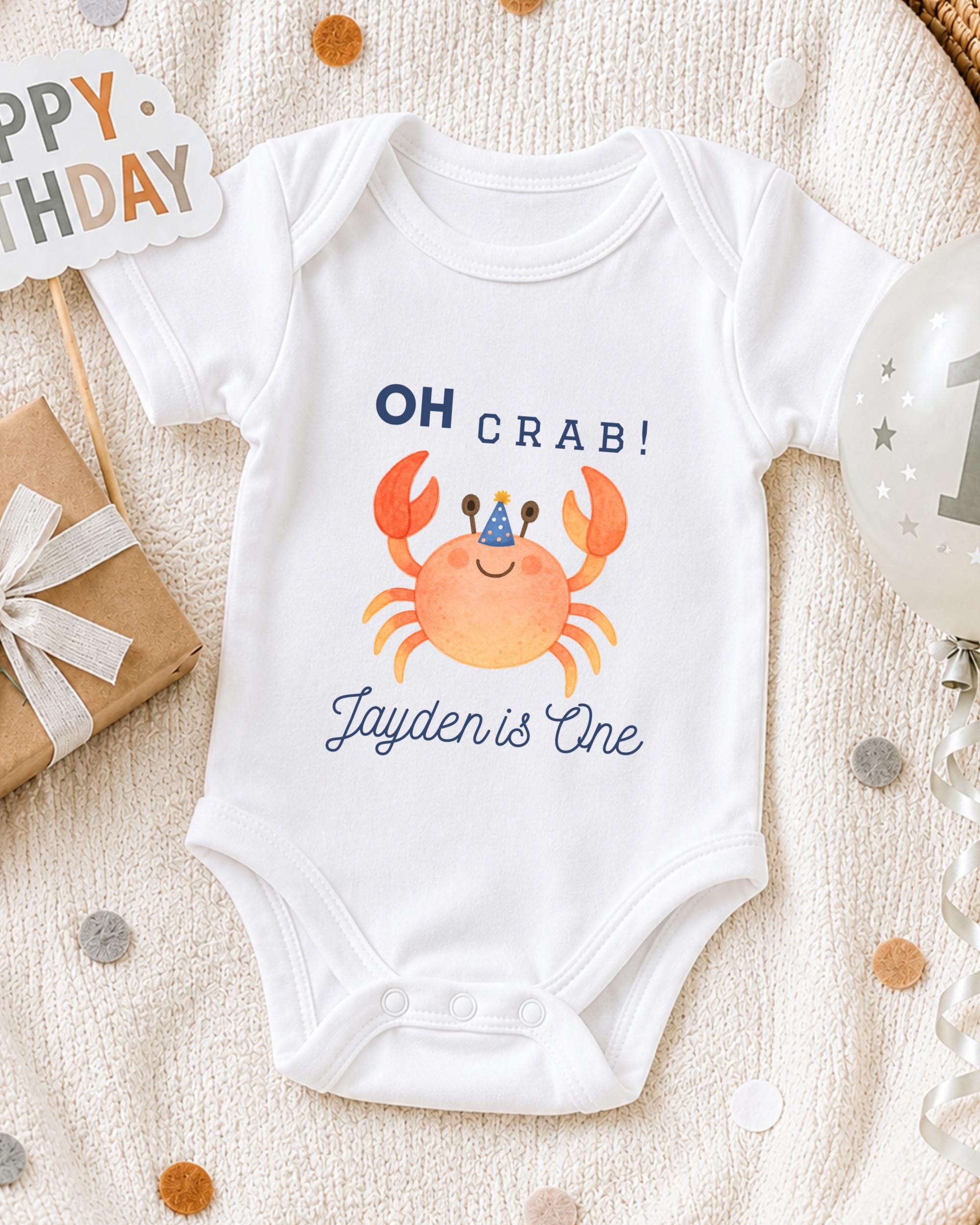 A white onesie with a crab design and the text "OH Crab! Jayden is One" printed on it, along with a birthday sign and a balloon.