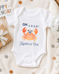 A white onesie with a crab design and the text "OH Crab! Jayden is One" printed on it, along with a birthday sign and a balloon.