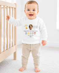 A smiling baby wearing a white sweatshirt with a personalized message and a photo of a man on it, standing in front of a wooden crib.