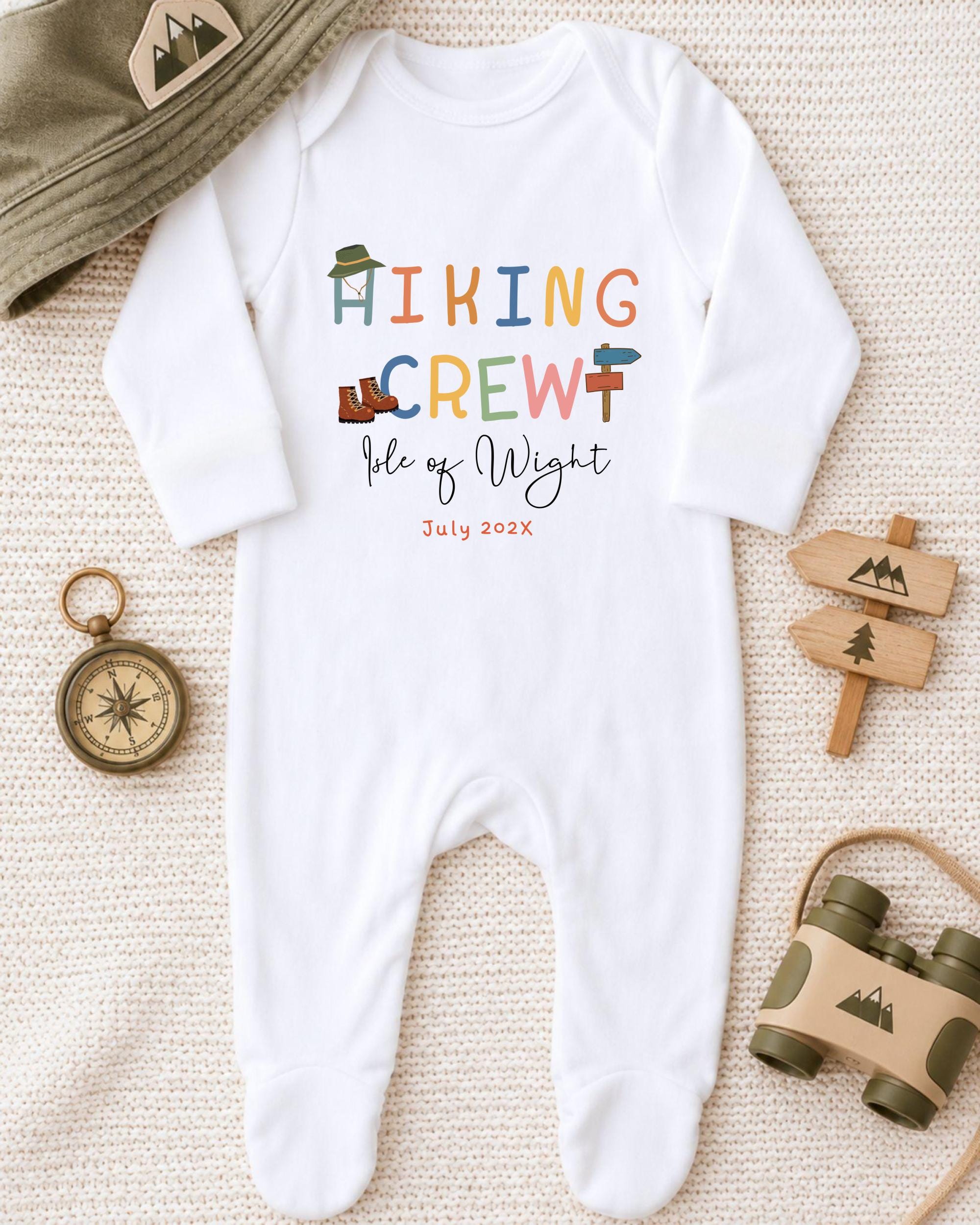 A baby onesie with the text "HIKING CREW" printed on it, along with a compass and binoculars, suggesting a theme related to outdoor activities or travel.