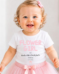 A smiling baby girl wearing a white t-shirt with pink text and a pink bow, standing in front of a white background.