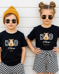 Two children wearing black t-shirts with a cartoon character and the text "BOO crew Tom" are standing against a white wall.