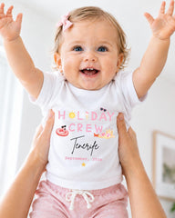 A smiling baby girl wearing a white t-shirt with the text "HOLIDAY CREW" and "Tenerife" printed on it, being held up by someone's arms.