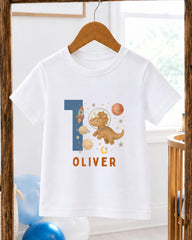 A white t-shirt with a dinosaur and space-themed design, along with the name "OLIVER" printed on it.