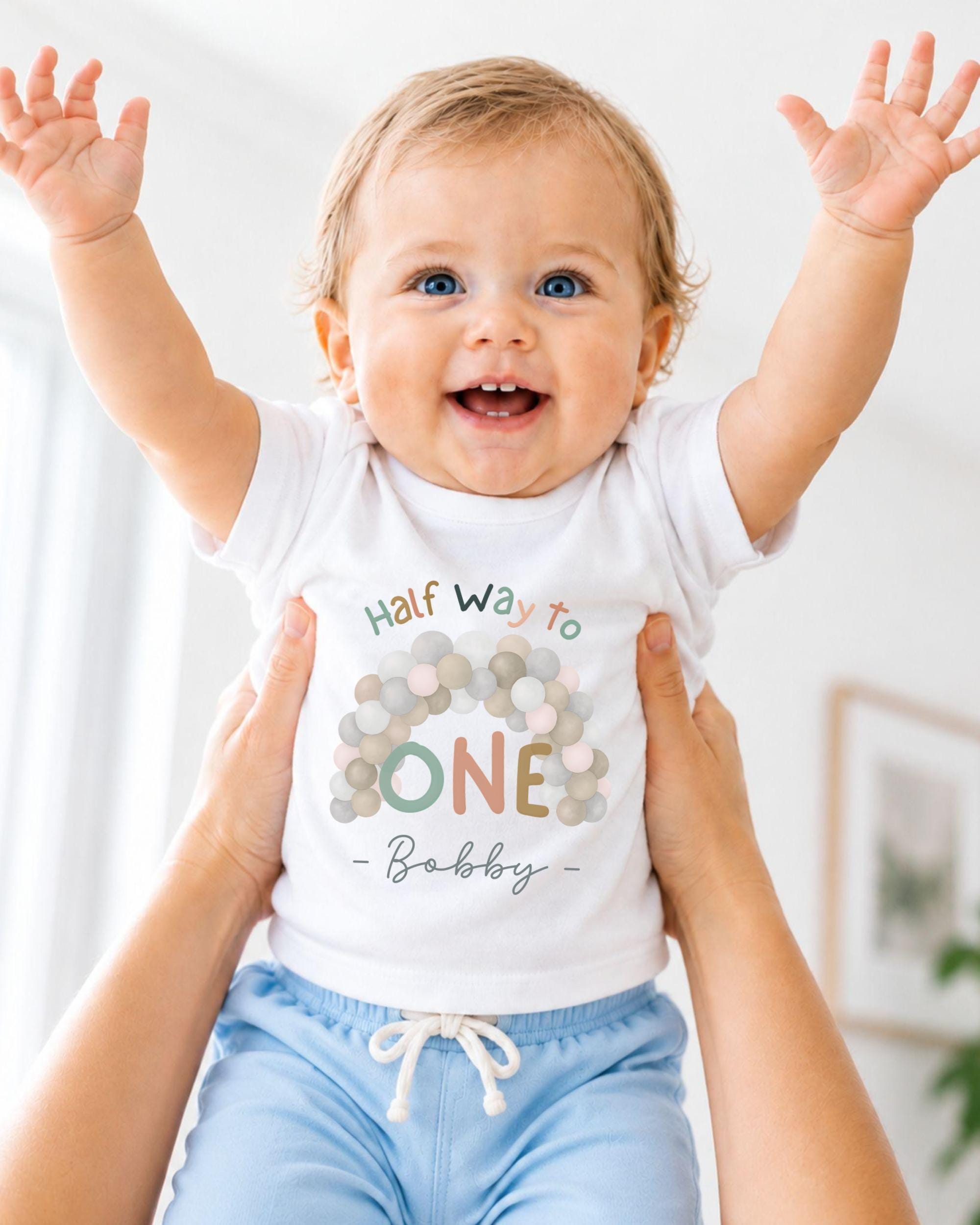 A smiling baby wearing a white t-shirt with a colorful design and the text "Half Way to One Bobby" is being held up by someone's arms.