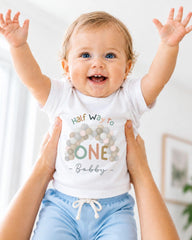 A smiling baby wearing a white t-shirt with a colorful design and the text "Half Way to One Bobby" is being held up by someone's arms.