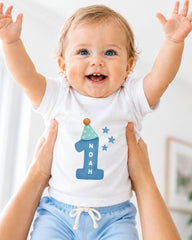 A smiling baby wearing a white onesie with a blue number one and stars, being held up by someone's arms.