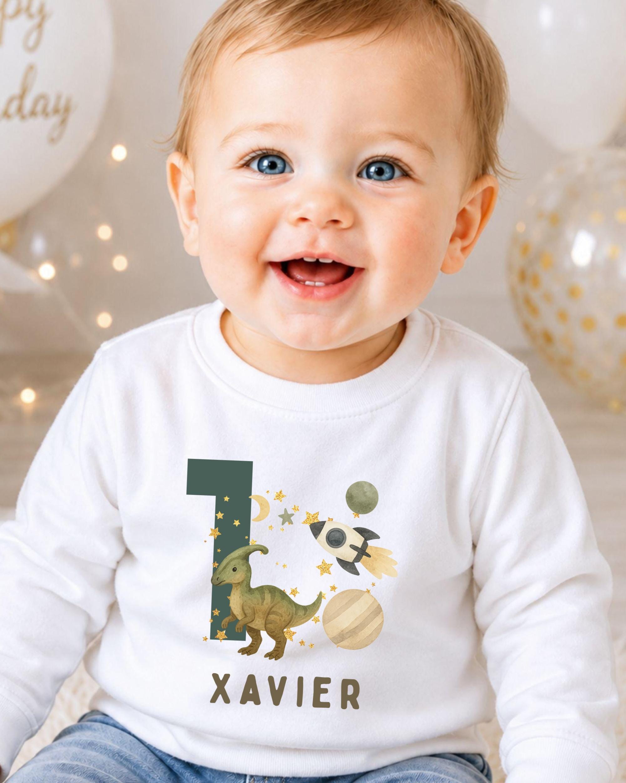 A smiling baby boy wearing a white shirt with a dinosaur and space-themed design, celebrating his first birthday.