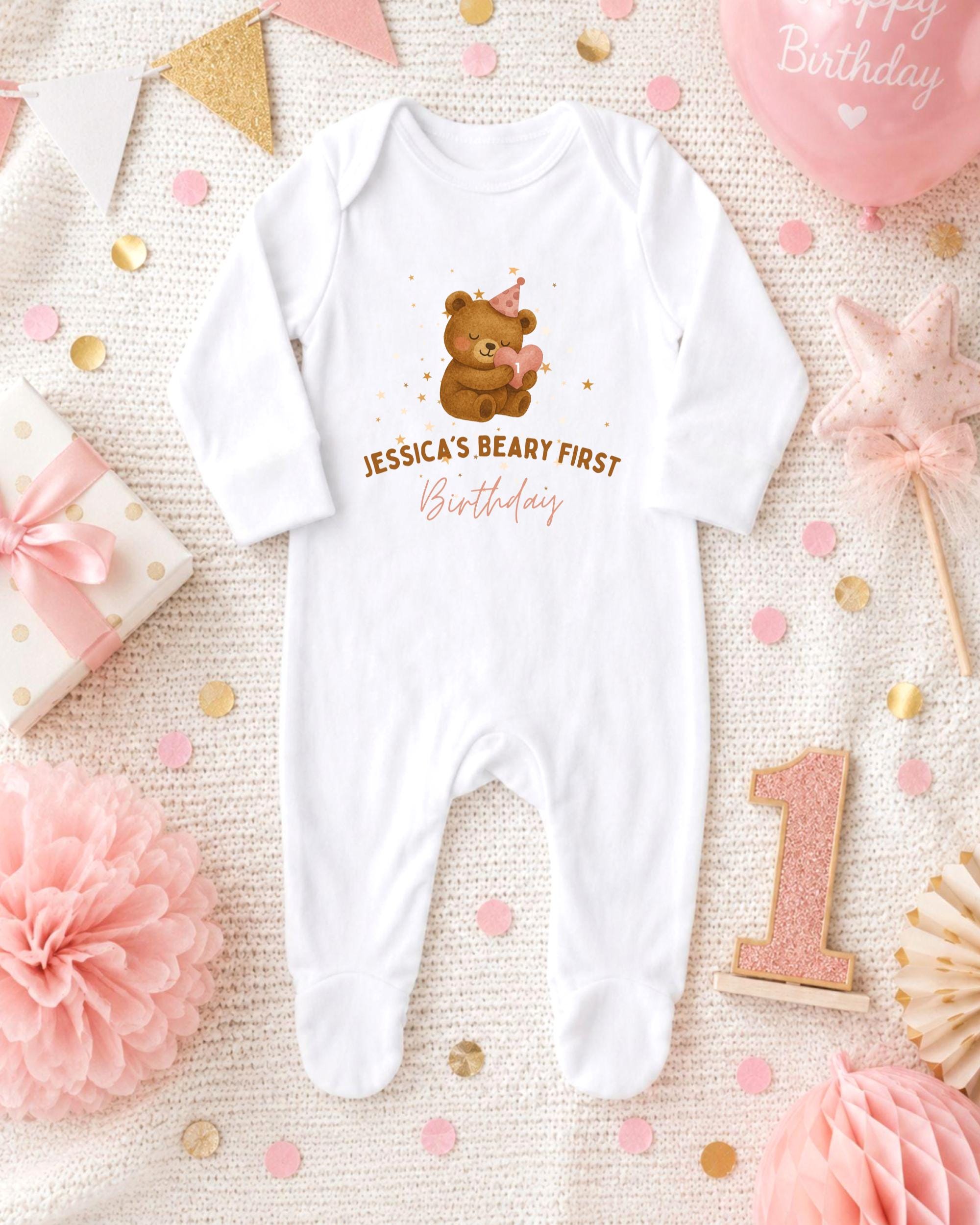 A white onesie with a teddy bear and the text " Jessica's first birthday" printed on it, surrounded by pink and gold confetti, a pink star, and a pink and gold birthday cake.