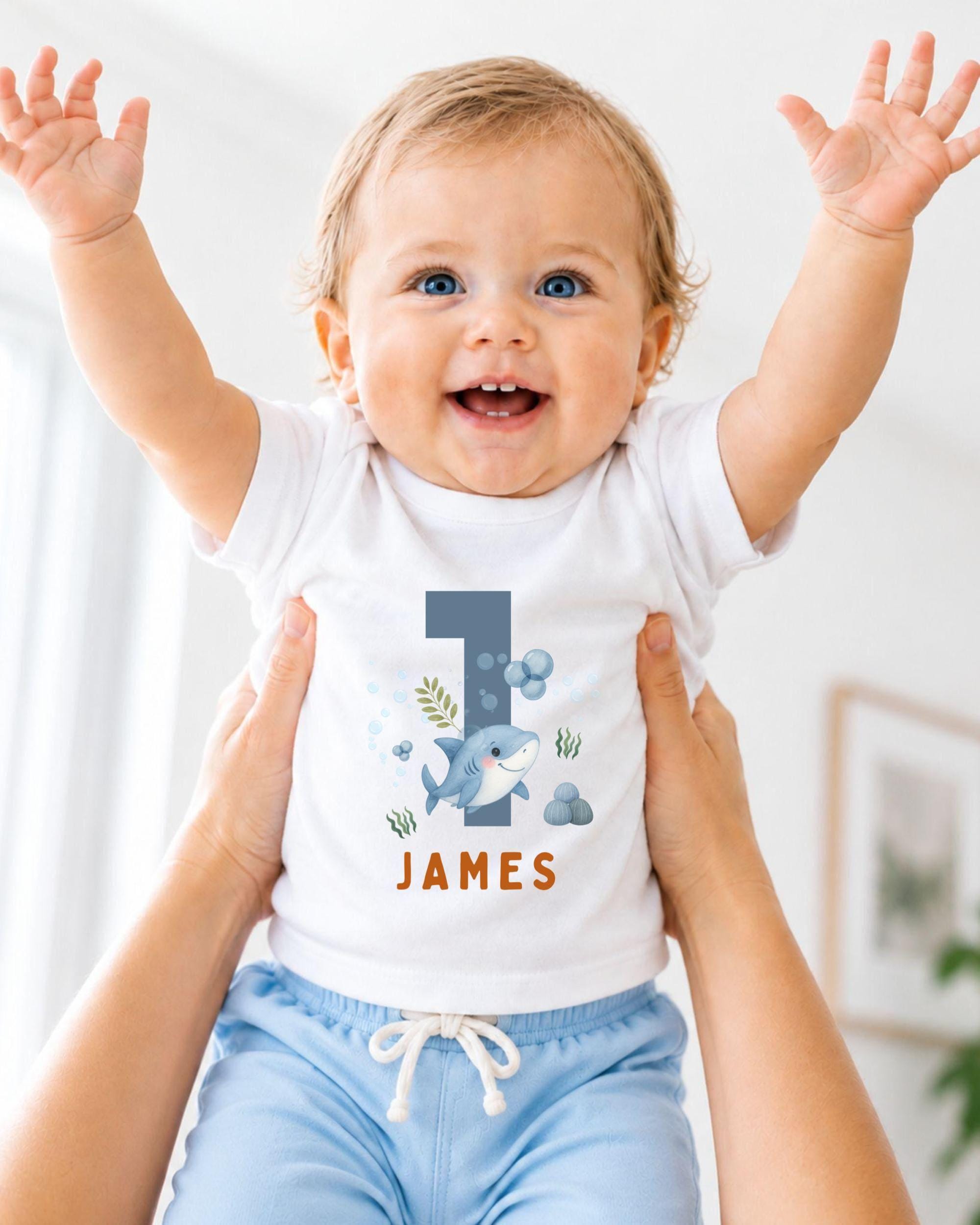 A smiling baby wearing a white shirt with a shark graphic and the name "James" printed on it, being held up by someone's arms.