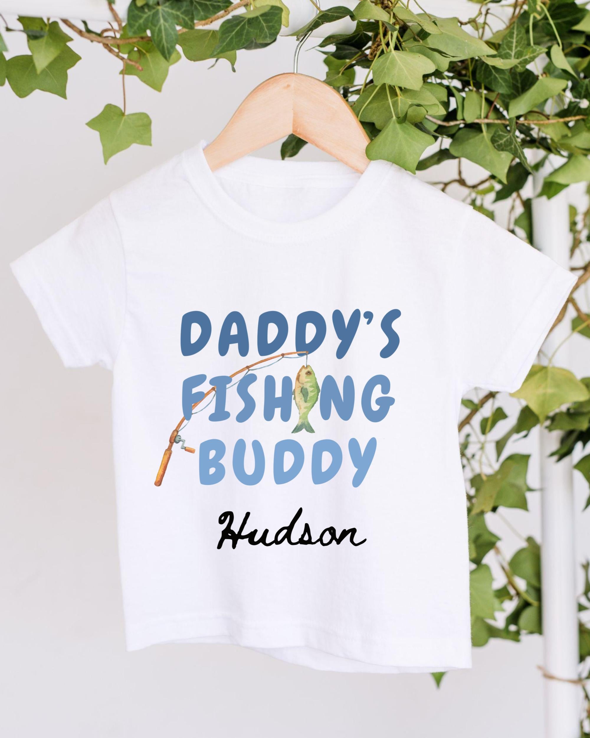 A white t-shirt with the text "DADDY'S FISHING BUDDY" printed on it, along with a fishing rod graphic. The t-shirt is hanging on a wooden hanger, and there are green leaves visible in the background.