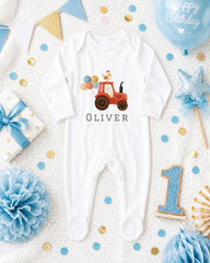 A white onesie with a tractor design, surrounded by birthday decorations such as a blue balloon, blue and gold star, blue and white polka dot gift box, blue and white pom pom, and blue and white confetti.