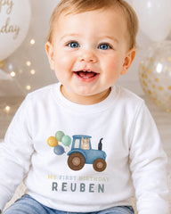 A smiling baby boy wearing a white shirt with a tractor and balloons printed on it, celebrating his first birthday.