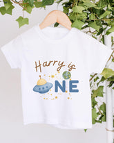 A white t-shirt with a graphic design featuring a spaceship and the text "Harry is one" printed on it.