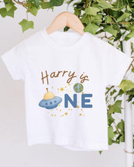 A white t-shirt with a graphic design featuring a spaceship and the text "Harry is one" printed on it.