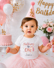 A young girl wearing a birthday hat and a white t-shirt with a unicorn design is sitting in front of a birthday cake and balloons.