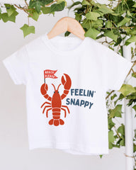 A white t-shirt with a red lobster graphic and text printed on it, hanging on a wooden hanger.