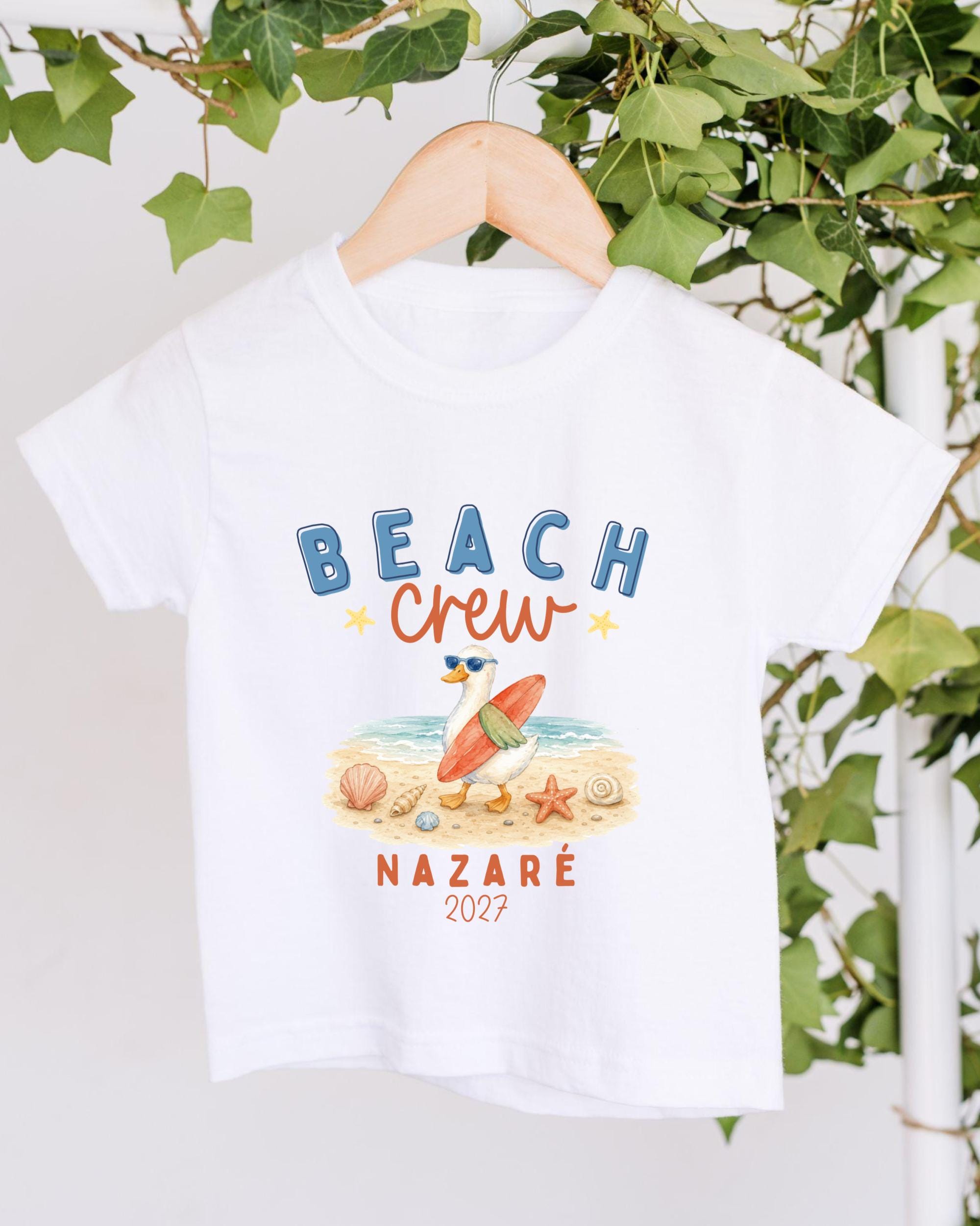 A white t-shirt with a beach-themed graphic and text printed on it, hanging on a wooden hanger.