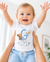A smiling baby wearing a white shirt with a letter "G" and a teddy bear graphic, being held up by someone's arms.