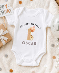 A white onesie with a giraffe and the text "My first birthday Oscar" printed on it, along with a "Happy Birthday" sign and a birthday balloon.