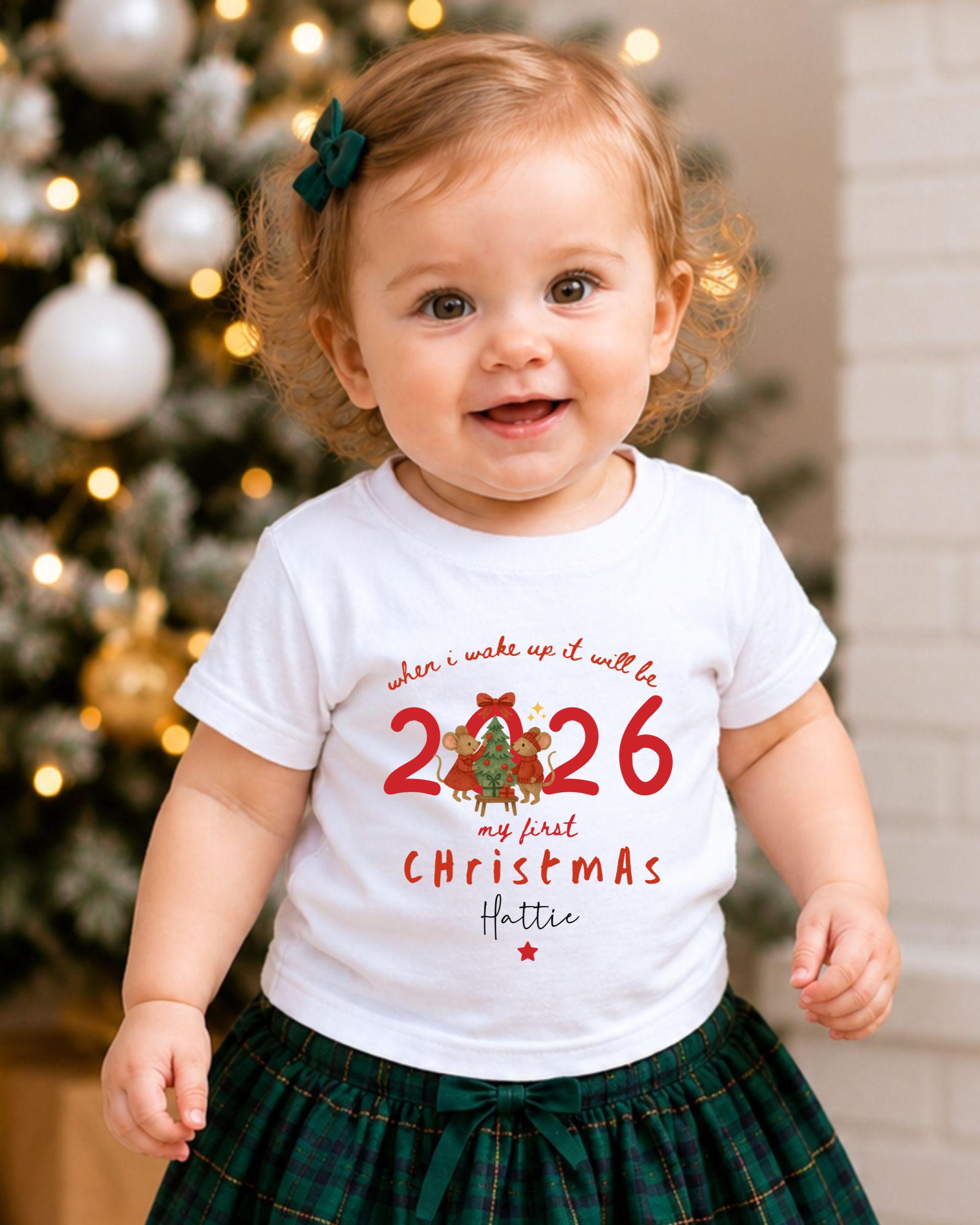 A smiling baby girl wearing a white t-shirt with a Christmas-themed design and a green bow in her hair, standing in front of a Christmas tree.